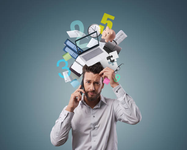 Stressed confused businessman having a phone call, he is surrounded by business items and office supplies: efficiency, planning and stress management concept